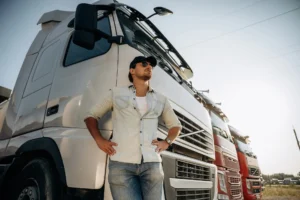 A male truck driver in a tan button-up shirt and jeans stands with his hands on his hips, leaning against the front of a white semi-truck. In the background, a line of other trucks is parked under a sunny sky.