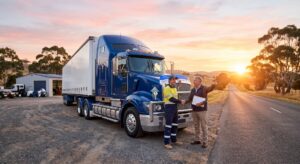 A detailed landscape photograph taken at sunrise, showing a large blue semi-truck with a trailer parked in a gravel depot. Two men are in the foreground, shaking hands in partnership. One man wears a high-visibility driver uniform, and the other is an older man holding business documents and pointing down a rural Australian road towards the golden sunlit horizon. An industrial workshop and eucalyptus trees form the background, signifying new beginnings and contracts