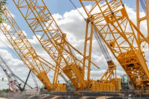 Lined up yellow crawler cranes with extended booms in auctioneer gravel lot for sale. Prospective buyers can apply for crane truck finance to purchase.